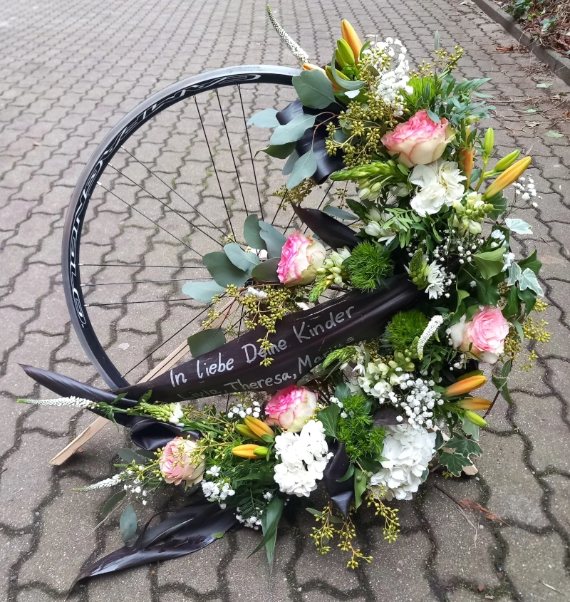 Trauergesteck auf einer Rennradfelge angebracht mit Lilien, Ornithogalum, Eucalypthus, Grasnelken, Löwenmaul, Rosen und Schleierkraut. Trauergesteck auf einer Rennradfelge angebracht mit Lilien, Ornithogalum, Eucalypthus, Grasnelken, Löwenmaul, Rosen und Schleierkraut.